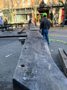 A large, weathered timber beam, part of a replica truss from the Notre Dame Cathedral, lies on a street in Ann Arbor, Michigan, during the Timber Framers Guild conference. A person wearing a hard hat is standing in the background.
