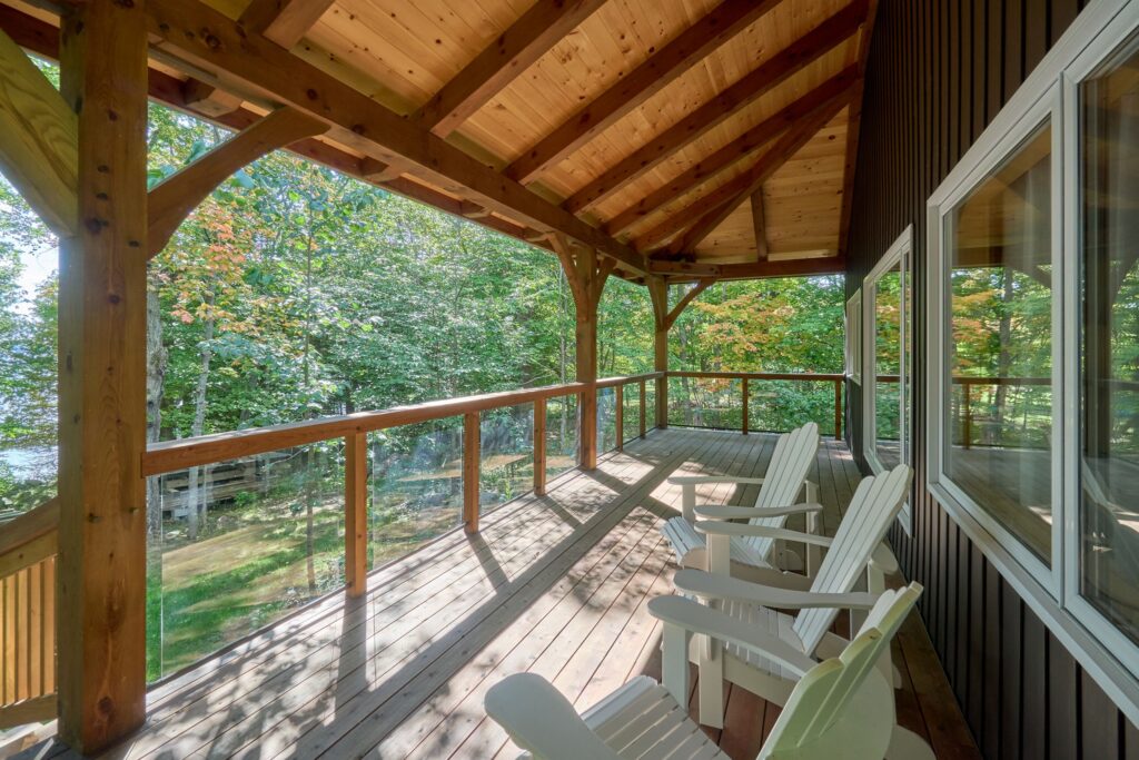 A view from the interior of the "Manitoulin Wrap-Around Porch" from the M'Chigeeng project. The porch features a timber frame roof, wooden flooring, and a modern glass railing. Four white Adirondack chairs face the forest and water view, with sunlight dappling the wooden deck.