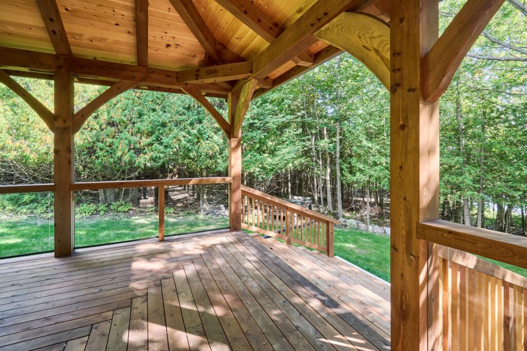 An interior perspective shot of the "Manitoulin Wrap-Around Porch" from the M'Chigeeng project. The image highlights the exposed timber frame roof structure with arched knee braces, tongue-and-groove cedar ceiling, wooden deck flooring, and the combination of traditional wooden stairs/railings with modern glass railings.