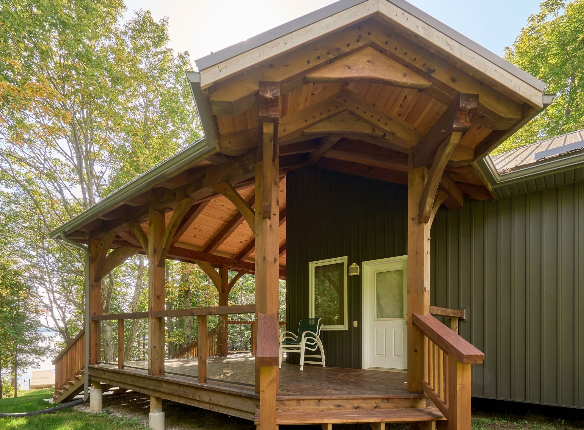An exterior view of the "Manitoulin Wrap-Around Porch" from the M'Chigeeng project. The porch features a natural wood finish, a detailed timber frame gable, a sloped access ramp, and stairs leading to a white entry door on a dark-sided house.