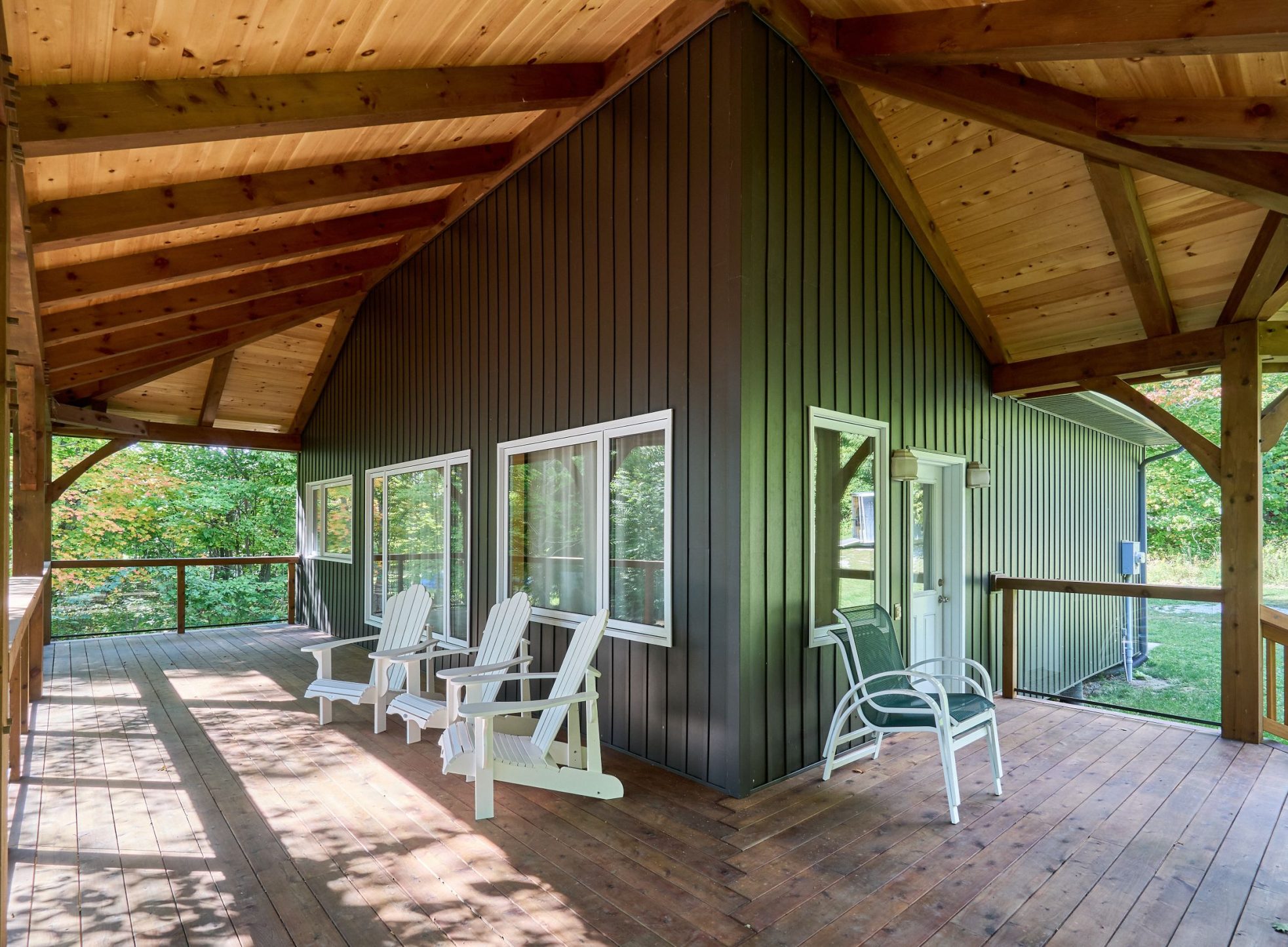 A wide-angle view of a covered timber frame porch from the M'Chigeeng project, featuring a wood-plank deck, exposed timber rafters and ceiling, and white Adirondack chairs facing a wooded area.